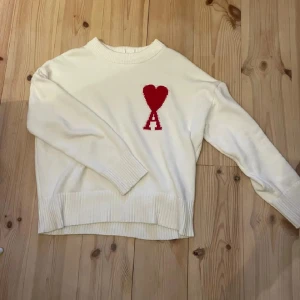 Vit stickad tröja från Ami stl M - Vit stickad tröja från Ami Alexandre Mattiussi med röd hjärt- och A-logga på bröstet. Tröjan har rund hals, ribbade muddar och lång ärm. Perfekt för dig som gillar stilrena detaljer och vill ha något som sticker ut lite extra.