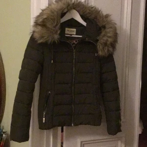 Svart pufferjacka med fuskpäls från Roco Baroco - Svart pufferjacka från Roco Baroco med fluffig fuskpäls på huvan, quiltad design och dragkedja framtill. Jackan har långa ärmar, två fickor med dragkedja och snygga detaljer. Perfekt för kalla dagar när du vill vara både varm och stilren.