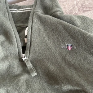Olivgrön half zip stickad tröja GANT - Olivgrön stickad tröja från GANT i superfin lammull. Tröjan har half zip med GANT-logga på dragkedjan och broderad logotyp på bröstet. Ribbad krage med vita ränder och långärmad modell. Perfekt för dig som gillar klassisk och chill stil.