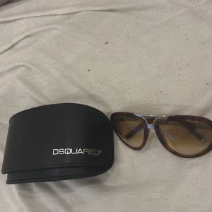 Bruna DSQUARED2 pilotglasögon - Snygga pilotglasögon från DSQUARED2 med bruna bågar och tonade linser. Metall-detaljer i silver på sidorna och bro. Kommer med original svart fodral. Perfekt för dig som vill ha en cool och klassisk look med designer vibe.