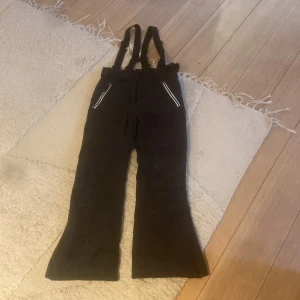 Svarta täckbyxor från Everest 140 - Svarta täckbyxor från Everest med hängslen och dragkedjor vid fickorna. Byxorna har breda ben och reflexdetaljer, perfekta för kalla dagar. Gjorda i slitstarkt syntetmaterial som står emot väder och vind.