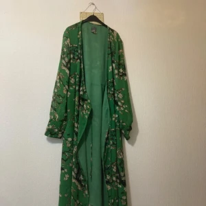 Grön blommig långklänning från ICHI - Grön långklänning från ICHI med blommigt mönster och transparent, lätt material. Klänningen har långa ärmar och knytband i midjan för en snygg siluett. Perfekt för dig som vill ha en flowy och chill look med extra detaljer. Köparen betalar för frakten