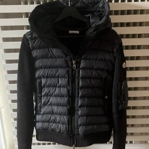 Svart Moncler pufferjacka med huva - Svart pufferjacka från Moncler med stickade ärmar och huva. Jackan har ribbade muddar, dragkedja fram och fickor med knappstängning. Moncler-logga på ärmen och en extra ficka med dragkedja på armen. Perfekt för dig som vill ha en snygg och varm jacka.