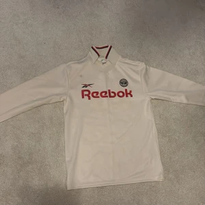 Vit långärmad fotbollströja Reebok - Snygg vit långärmad fotbollströja från Reebok med röd logga och klubbmärke på bröstet. Tröjan har klassisk krage med röda och svarta detaljer. Perfekt för dig som gillar retrostil och sportmode.