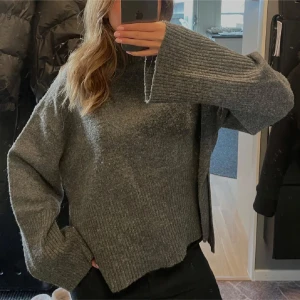 Grå stickad polotröja med slits - Mysig grå stickad polotröja med breda ribbade ärmslut och hög hals. Tröjan har en avslappnad passform och cool slits på sidan som ger en extra detalj. Perfekt för lager-på-lager och chill dagar.