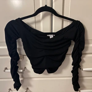 Svart croppad topp från AMISU - Säljer en svart croppad topp från AMISU med långa, rynkade ärmar och bred urringning. Toppen har en tight passform och coola draperade detaljer framtill. Perfekt för dig som gillar trendiga och stilrena plagg.