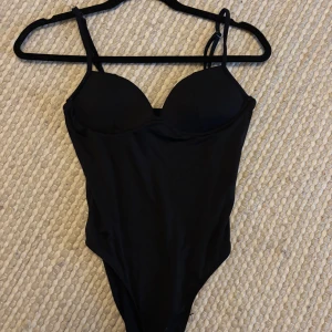 Svart body med bygel och smala band - Snygg svart body med vadderade kupor och bygel för extra stöd. Modellen har smala axelband och en klassisk, stilren look. Materialet känns mjukt och följsamt mot kroppen.