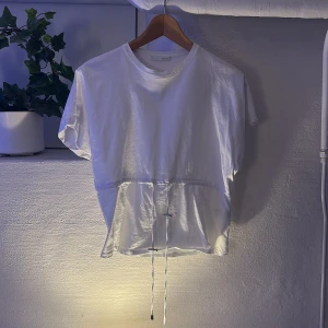 Vit kortärmad topp med snörning från Zara - Säljer en vit kortärmad topp från Zara med rund hals och cool snörning i midjan som ger en snygg siluett. Toppen är gjord i mjuk bomull och har en enkel, clean look som funkar till många stilar. Perfekt för dig som gillar minimalistiskt och fräscht.