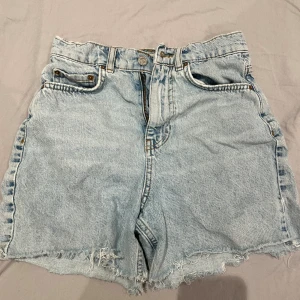 Ljusblå denimshorts med fransig kant - Ljusblå denimshorts från Perfect Jeans med hög midja och rå, fransig kant nertill. Klassisk femficksmodell med dragkedja och knapp framtill. Perfekta för sommardagar och enkel att matcha med allt.