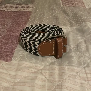 Svartvitt flätat bälte med läderdetaljer - Snyggt flätat bälte i svart och vitt med bruna läderdetaljer och silverfärgat spänne. Perfekt accessoar för att lyfta din outfit med en modern touch. Bältet har ett randigt mönster och är gjort i syntetmaterial med läderdetaljer.