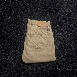 Beige chinos från Atelier Gardeur 34/34 - Snygga beige chinos från Atelier Gardeur i storlek 34/34. Klassisk modell med bakficka och diskret logopatch i läder vid linningen. Byxorna är tillverkade i mjuk bomull och har en stilren look som passar till många outfits.