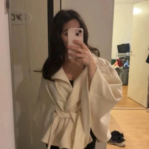 Vit poncho jacka  - Vit creme poncho med bälte i midjan. Jackan har vida ärmar och en mjuk, slät yta som ger ett elegant intryck. Perfekt för lager-på-lager och enkel att matcha med olika outfits.