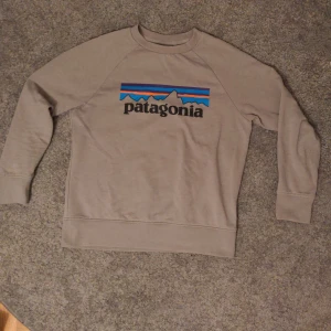 Grå/Beige Patagonia sweatshirt i bomull storlek 142 - Tröjan är i storlek 142/M i barnstorlekar. Säljer en grå-beige sweatshirt från Patagonia i ekologisk bomull. Tröjan har klassisk rund hals, lång ärm och stor Patagonia-logga med detaljer framtill.