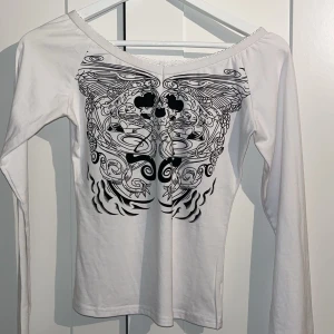 Vit långärmad topp med svart tryck - Säljer en vit långärmad topp med ett stort, detaljerat svart tryck framtill som har ett symmetriskt och konstnärligt mönster. Toppen har en bred halsringning med liten spetskant och är figurnära i modellen. Perfekt för dig som gillar unika prints!
