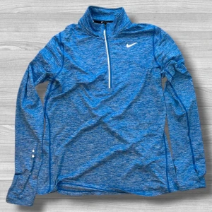 Nike blå Dri-Fit half zip träningströja - Nike Dri-Fit långärmad träningströja i melerad blå färg med vit logga på bröstet. Tröjan har half zip-dragkedja framtill och är tillverkad i ett lätt, syntetiskt material som andas. Perfekt för löpning eller gym. Snygg och sportig look.