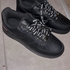 Nike Air Force 1 x Supreme svarta sneakers - Svarta Nike Air Force 1 sneakers i samarbete med Supreme. Skorna har Supreme-logga på sidan och svarta snören med vit Supreme-text. Ovandel i skinn, perforerad tå och platt sula. Klassisk rund tå och lågt skaft. Perfekt för dig som gillar streetwear och exklusiva collabs. Nyskick 
