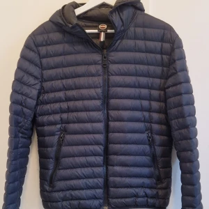 Mörkblå Colmar dunjacka - Säljer en mörkblå dunjacka från Colmar med huva och två sidofickor med dragkedja. Jackan har ett quiltat mönster och Colmar-logga på ärmen. Sparsamt använd så mycket fint skick. Storlek 48 (M) men ganska liten i storlek så mer en S.