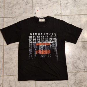 Maison Margiela svart t-shirt med print - Svart t-shirt från Maison Martin Margiela med ikoniskt sifferprint och texten 'Maison Martin Margiela PARIS' framtill. Trycket har en cool, sliten effekt i vitt och orange. Lite oversized passform med rund hals och korta ärmar. Perfekt statement-plagg för dig som gillar designer. Storlek S. MM tryck på framsidan och den ikoniska 'stitch' loggan på baksidan. Denna har fått en sjukt snygg sunfade på baksidan runt kragen men går att tvätta bort vid första tvätt. Hör av er vid minsta fråga! 🤍🙌