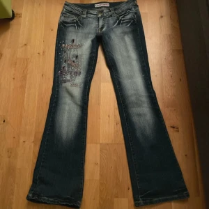 Blå bootcut jeans med broderi - Snygga blå bootcut jeans från Porta Fortuna med coolt broderi och paljettdetaljer på vänster lår. Jeansen har klassisk femficksdesign och slitningar för en trendig look. Perfekt för dig som gillar unik stil och detaljer.