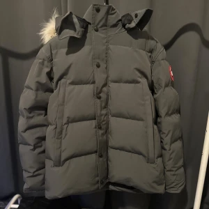 Grå Canada Goose Wyndham Jacka med Äkta päls - Snygg och varm dunjacka från Canada Goose i grå färg med äkta päls på huvan. Klassisk pufferstil med dragkedja och knappar framtill, samt avtagbar luva/päls. Ikonisk logga på ärmen och kommer med alla tillbehör.  Perfekt för kalla vinterdagar – håller värmen samtidigt som den ser exklusiv ut. Pris kan diskuteras vid snabb affär. Fraktas endast.