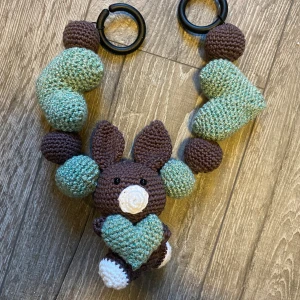 Handvirkad barnvagnsleksak med kanin - Charmig handvirkad barnvagnsleksak med söt kanin, hjärtan och bollar i grönt och brunt. Fästs enkelt med plastclips. Mjukt garn, inga synliga defekter eller slitage. Perfekt för att stimulera och underhålla små barn under promenader.