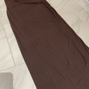 Brun långklänning från lager 157 - Säljer en stilren brun långkjol från lager 157 i storlek XS. Kjolen har en enkel och rak passform men ändå följer kroppsformen och är tillverkad i ett mjukt och stretchigt material. Den går ner hela vägen till fötterna. Helt ny med prislapp aldrig använt