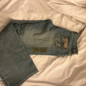 Blå jeans med nitar från TARA - Säljer ett par blå jeans från TARA i 100% bomull. Jeansen har utsvängda ben, coola nitar och stenar på bakfickan samt ett tryck i guld på ena benet. Perfekt för dig som vill sticka ut med detaljer och streetkänsla.