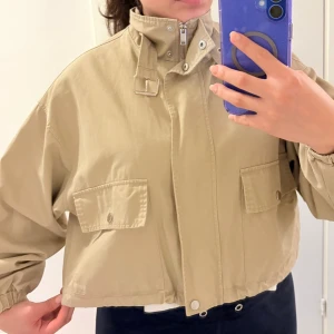 Beige croppad vindjacka med hög krage - Snygg beige vindjacka med croppad passform, hög krage med spänne och dragkedja framtill. Jackan har två stora fickor med lock och tryckknappar samt elastiska ärmslut. Perfekt för dig som gillar en avslappnad och trendig stil.