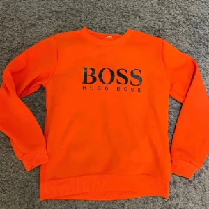 Orange sweatshirt från Hugo Boss - Säljer en snygg orange sweatshirt från Hugo Boss med svart logga framtill. Tröjan har rund halsringning och långa ärmar med ribbade muddar. Perfekt för dig som vill sticka ut med färg i din outfit.