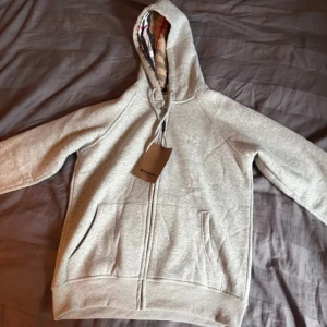 Grå hoodie från Burberry med dragkedja - Snygg grå hoodie från Burberry med klassiskt rutigt foder i huvan. Tröjan har dragkedja framtill, två fickor och snörning vid huvan. Perfekt för dig som gillar stilrena detaljer och vill ha något som sticker ut med det ikoniska Burberry-mönstret.