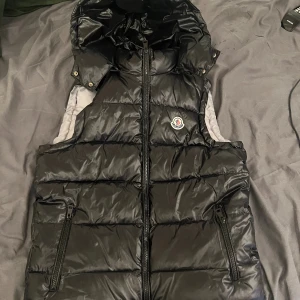 Svart dunväst från Moncler med huva - Snygg svart dunväst från Moncler med glansig finish, avtagbar huva och dragkedja framtill. Västen har två fickor med dragkedja och klassisk Moncler-logga på bröstet. Insidan är ljusgrå och har en innerficka med dragkedja.