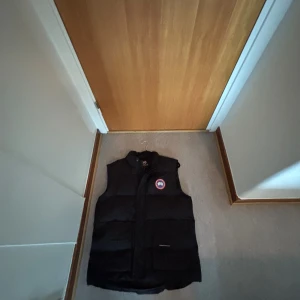 Svart dunväst från Canada Goose - Svart dunväst från Canada Goose med klassisk logga på bröstet. Västen har hög krage, två stora fickor framtill och stängs med både dragkedja och knappar. Perfekt för lager-på-lager och riktigt snygg till streetwear.