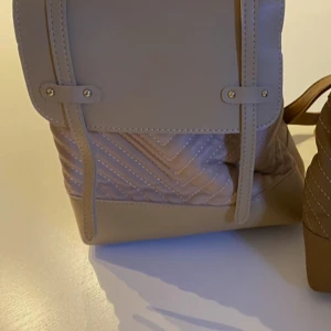 Beige quiltad ryggsäck i skinnimitation - Snygg beige ryggsäck i skinnimitation med quiltade detaljer och justerbara axelband. Väskan har en stilren design med dekorativa sömmar och metallspännen. Perfekt för dig som vill ha en trendig och praktisk accessoar till vardagen.
