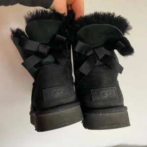 Svarta UGG boots med rosett - Mysiga svarta UGG boots i mocka med fluffig insida och dekorativa svarta rosetter baktill. Skorna har rund tå, platt sula och är perfekta för kalla dagar. Klassisk UGG-logga på hälen och en stilren look som passar till många outfits.