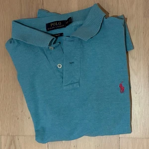 Blå pikétröja från Polo Ralph Lauren - Snygg blå pikétröja från Polo Ralph Lauren med klassisk krage och två knappar framtill. Tröjan har korta ärmar och den ikoniska rosa broderade loggan på bröstet. Perfekt för dig som gillar stilrena och tidlösa plagg.