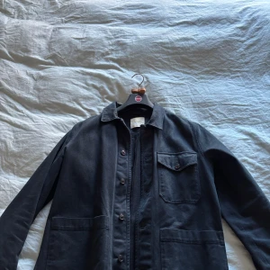 Mörkblå overshirt från A Day's March - Stilren svart overshirt från A Day's March i bomull. Klassisk krage, knäppning framtill med knappar och tre fickor – två stora nedtill och en bröstficka med lock. 7/10 skick då ena knappen på ärmen saknas men annars är jackan superfin! 