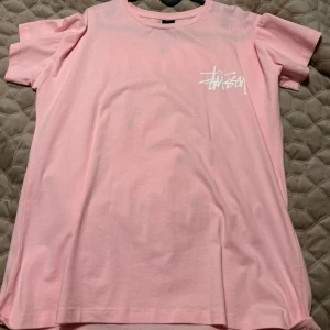 Rosa Stüssy t-shirt med tärningsprint - Säljer en rosa t-shirt från Stüssy med vit logga på bröstet och stor print på ryggen med två rosa tärningar och Stüssy-logga ovanför. T-shirten är i mjuk bomull och har klassisk passform med korta ärmar och rund hals.