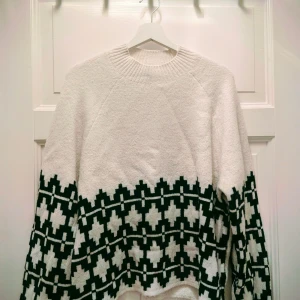 Vit stickad tröja med svart mönster - Säljer en vit stickad tröja med svart grafiskt mönster längs nederdelen och ärmarna. Tröjan har rund hals och långa ärmar, perfekt för att hålla sig varm och snygg under kyliga dagar. Mjuk och skön känsla, passar dig som gillar stilrena detaljer. Divided, storlek M medium, dam