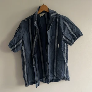 Blå randig skjorta från Yoke - Snygg kortärmad skjorta från Yoke i blå nyanser med vertikala ränder i olika toner av blått och grått. Skjortan har klassisk krage, bröstficka och knappar framtill. 