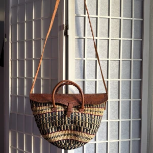 Randig axelväska med skinn och flätad design - Snygg axelväska med handtag i brunt skinn och lock med dekorativa mönster. Väskan är flätad i naturmaterial med svarta, beige och röda ränder. Unik form och coolt mönster, perfekt för dig som gillar boho-vibe och vill sticka ut.