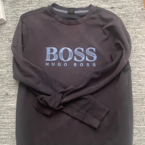 Svart långärmad tröja Hugo Boss S - Svart långärmad tröja från Hugo Boss i storlek S. Tröjan har ett stort blått BOSS-tryck på bröstet och rund hals. Mjuk bomullskänsla och klassisk passform, perfekt för dig som gillar stilrena logotröjor.