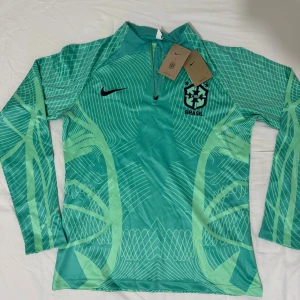 Grön Brasilien långärmad Nike fotbollströja - Snygg långärmad Brasilien fotbollströja från Nike med Dri-FIT material. Tröjan är grön med ljusgröna grafiska mönster och har halv dragkedja framtill. Brasil-logga och Nike Swoosh på bröstet. Perfekt för träning eller match!