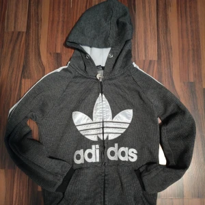 Svart Adidas zip hoodie  - Svart hoodie från Adidas med vit stor trefoil-logga framtill och vita ränder längs ärmarna. Tröjan har dragkedja, huva och är tillverkad i 100% bomull. Perfekt för en sportig och avslappnad stil. Storlek 134-140 barn.
