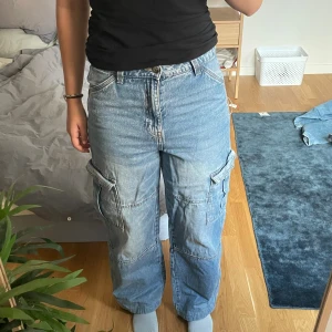 Blå cargobyxor i jeans från H&M Divided - Säljer ett par blå cargobyxor i jeansmaterial från H&M Divided, storlek 36. Byxorna har flera stora fickor på sidorna, raka ben och en avslappnad passform. De är uppsydda för att passa någon som är 165cm. Klassisk ljusblå denim med synliga sömmar och bälteshällor.🥰🥰Pris kan alltid diskuteras🥰💛