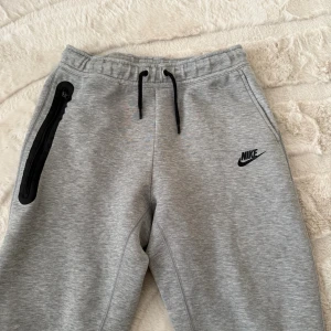 Grå mjukisbyxor Nike tech  - Grå mjukisbyxor från Nike med svart logga på benet, svarta dragsnören i midjan och en praktisk dragkedjeficka på sidan. Tillverkade i mjukt bomullsmaterial för maximal komfort. Perfekta för chill eller träning.