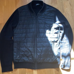 Svart cardigan från Barbour International - Svart cardigan från Barbour International med quiltad front och släta ärmar. Jackan har ribbade muddar, två fickor med dragkedja och en snygg patch på ärmen. Perfekt för dig som gillar stilren och sportig look. Passar dig runt 180, säljs inte nu längre. 