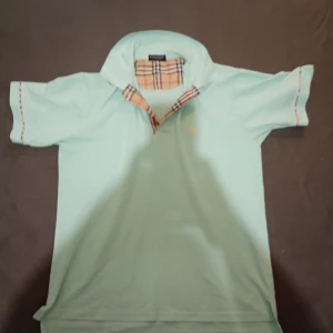 Mintgrön  skjorta med rutig krage - Fräsch mintgrön skjorta med klassisk rutig krage i beige, svart och rött. Skjortan har korta ärmar och en liten broderad logga på bröstet. Perfekt för dig som gillar stilrena detaljer och ikoniska mönster.