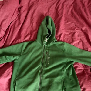 Grön hoodie från Sail Racing XL - Grön hoodie från Sail Racing i storlek XL. Tröjan har hel dragkedja, huva och en bröstficka med dragkedja. Broderad logga på bröstet och stor text på ryggen. Mjuk fleece på insidan för extra komfort.