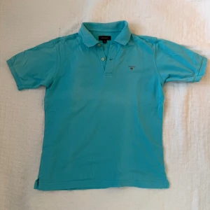 Turkos pikétröja från GANT ungdom 146/152 - Snygg turkos pikétröja från GANT med klassisk krage och två knappar framtill. Tröjan har korta ärmar och GANT-logga broderad på bröstet. Liten tendens till fläck, se bild. Stl ungdom  10-12 år 146/152