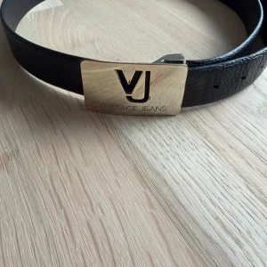 Svart Versace Jeans bälte i skinn - Snyggt svart bälte från Versace Jeans med bred design och exklusivt guldigt spänne med VJ-logga. Tillverkat i äkta skinn och har en stilren look som lyfter din outfit. Bältet är tillverkat i Italien och har märkets signaturdetaljer. Bältet är äkta och använts 3 gånger. 